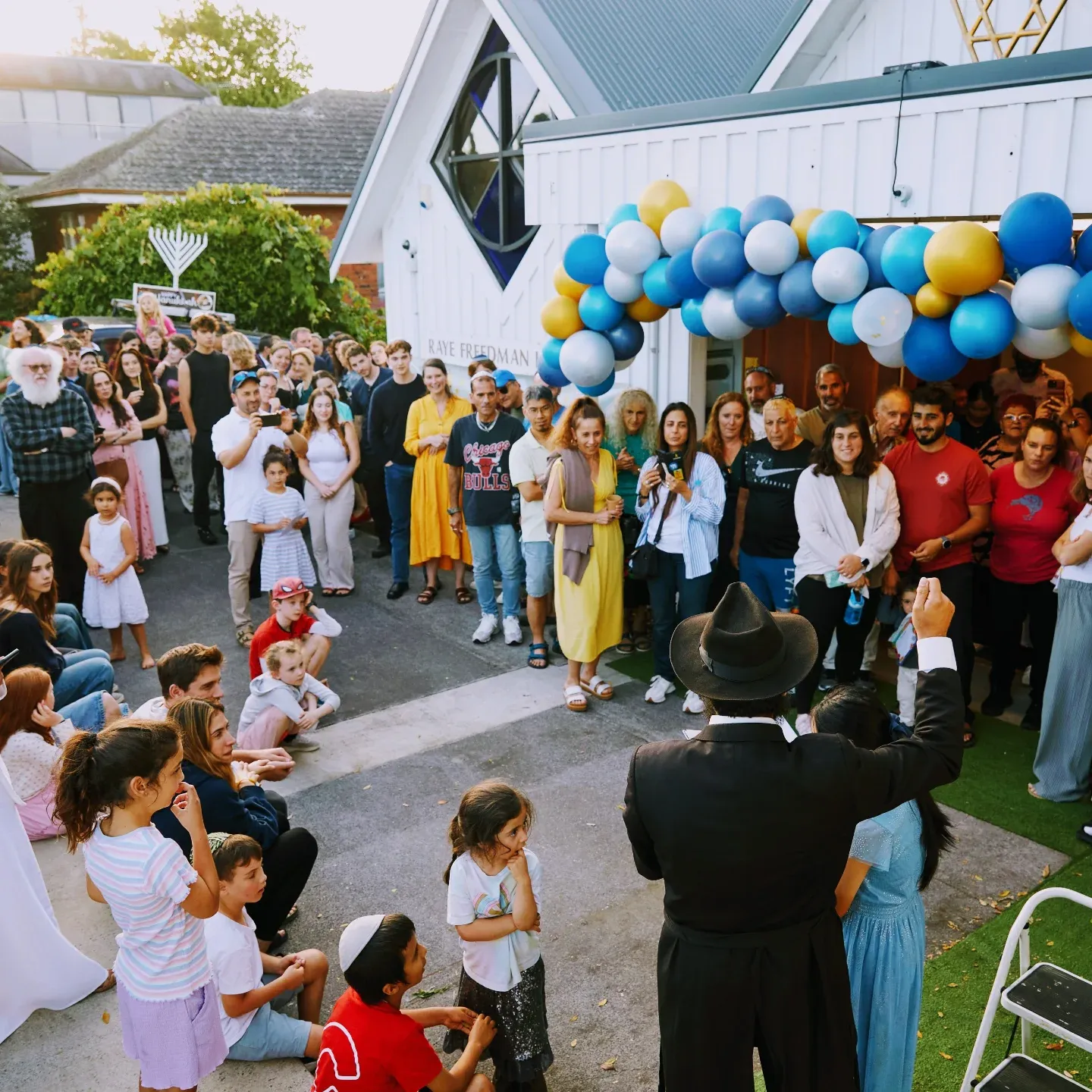 A large crowd of families gathered outside the Raye Freedman Library at 788 Remuera Road beneath blue and yellow balloons, a rabbi addressing them