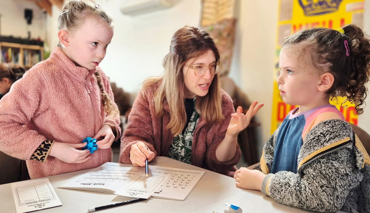 A teacher explaining Hebrew letters to two young girls at a Hebrew School table